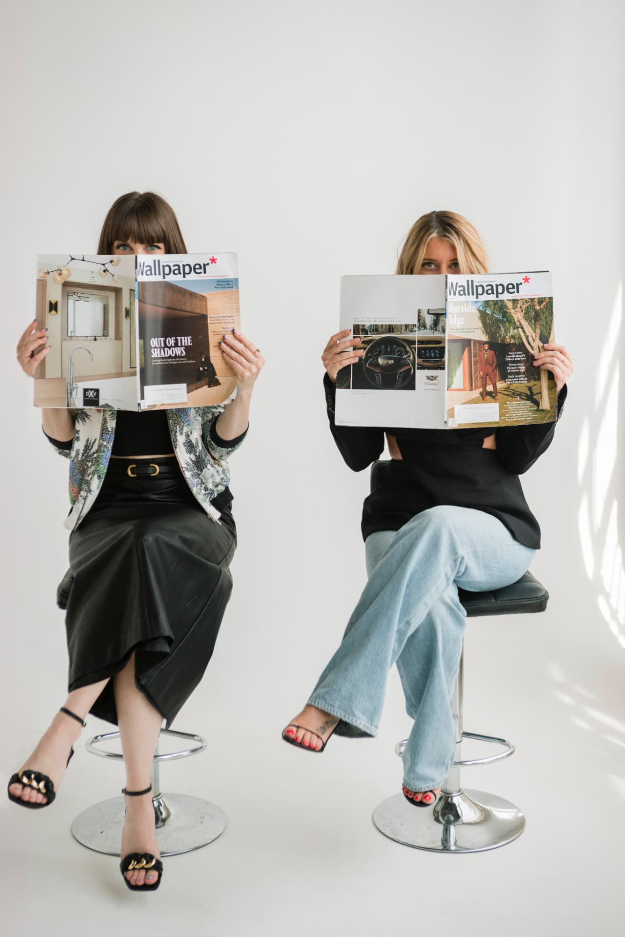 Two women seated on stools holding open magazines in front of their faces