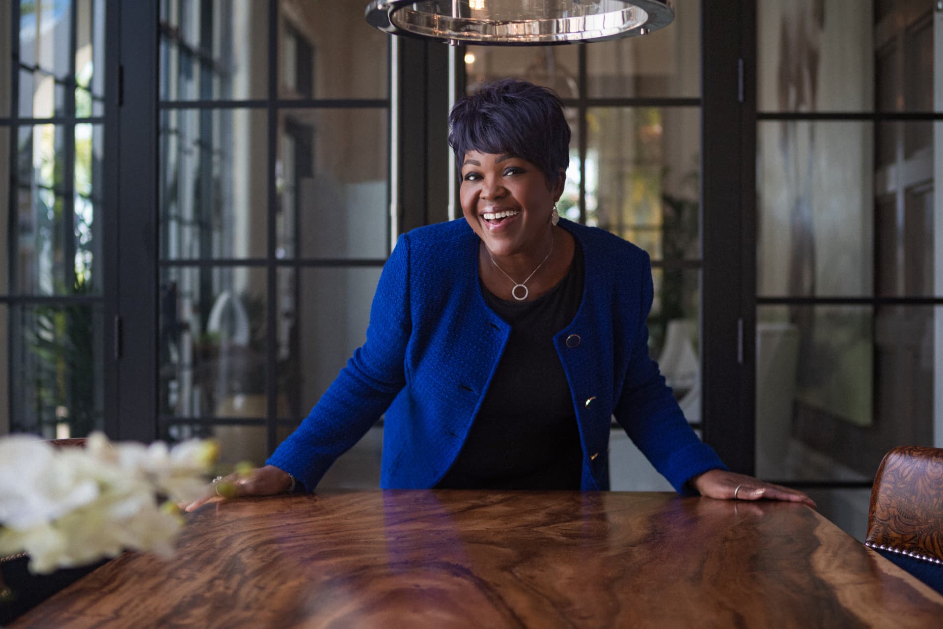 A woman laughing warmly at a wooden dining table, wearing a cobalt blue jacket